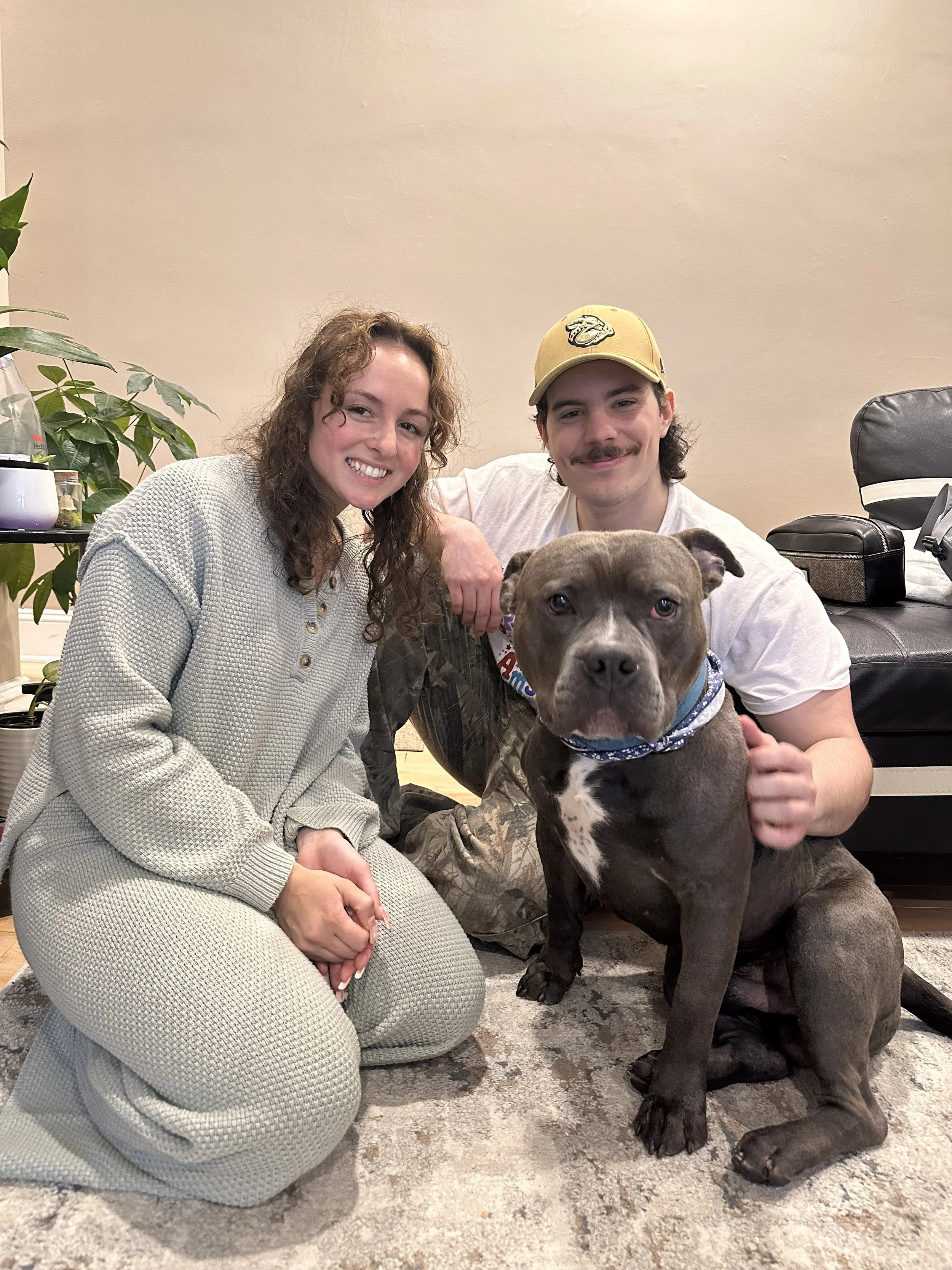 A woman, a man, and a large dog sitting on a rug inside a living room. The woman has curly hair and is wearing a gray knit outfit, and the man is wearing a white shirt, a yellow cap, and camouflage pants. Both are smiling at the camera.