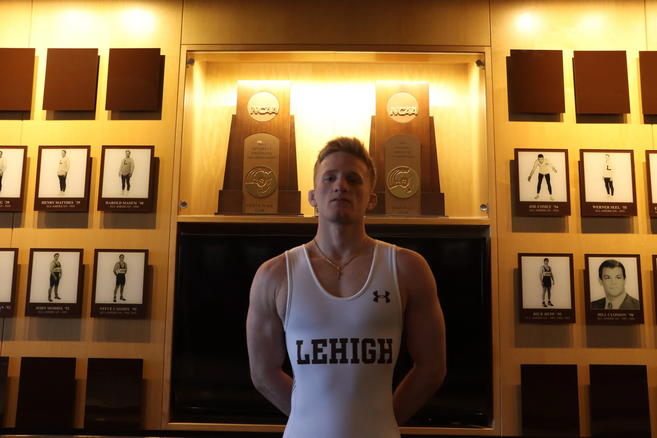 A young man in a white athletic jersey with 'LEHIGH' written on it, standing in front of a wall displaying framed black-and-white photos of athletes and trophies.