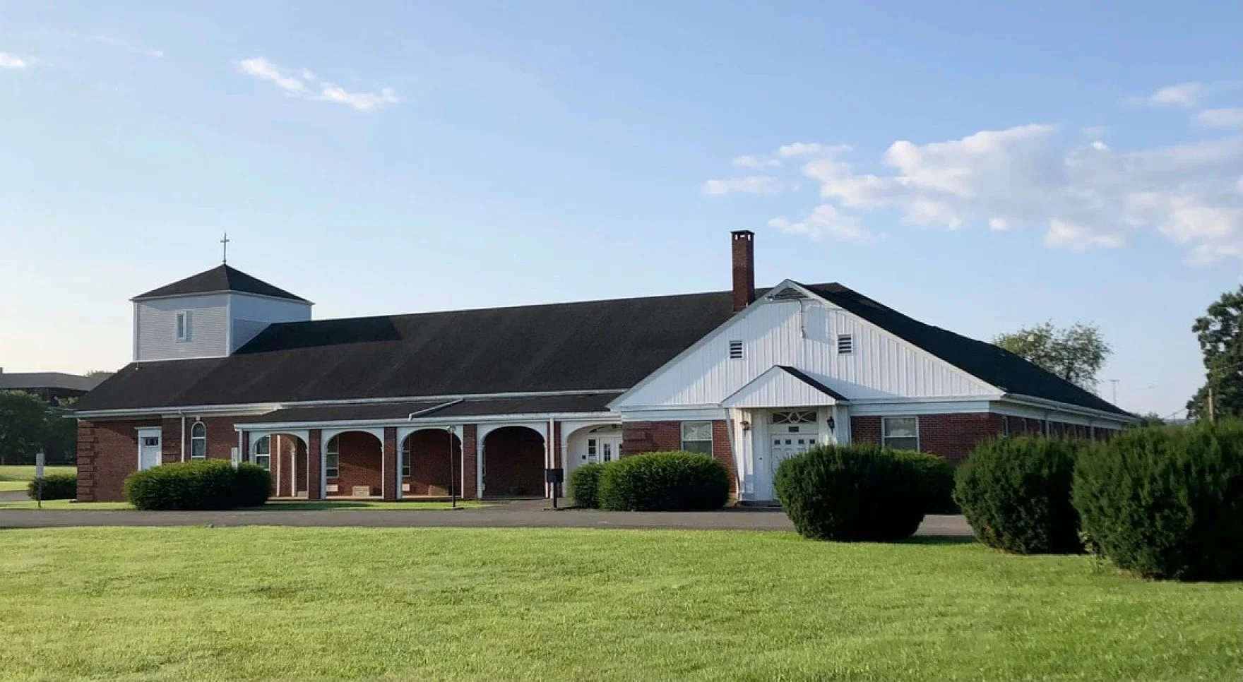 A brick church with a white steeple and a porch, surrounded by well-maintained bushes and grass, under a partly cloudy sky.