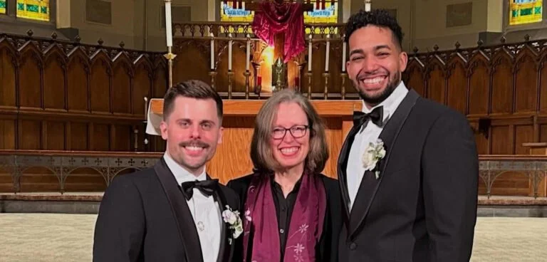 Two men in tuxedos with bow ties and a woman in a black dress and glasses standing inside a church with stained glass windows and wooden pews. They are smiling.