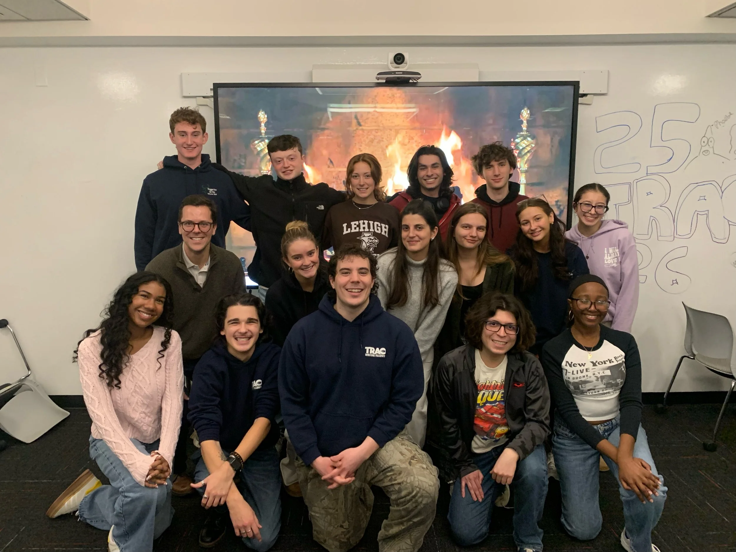 Group of diverse young students and a teacher posing together in a classroom, with a whiteboard and TV screen behind them displaying a fireplace.
