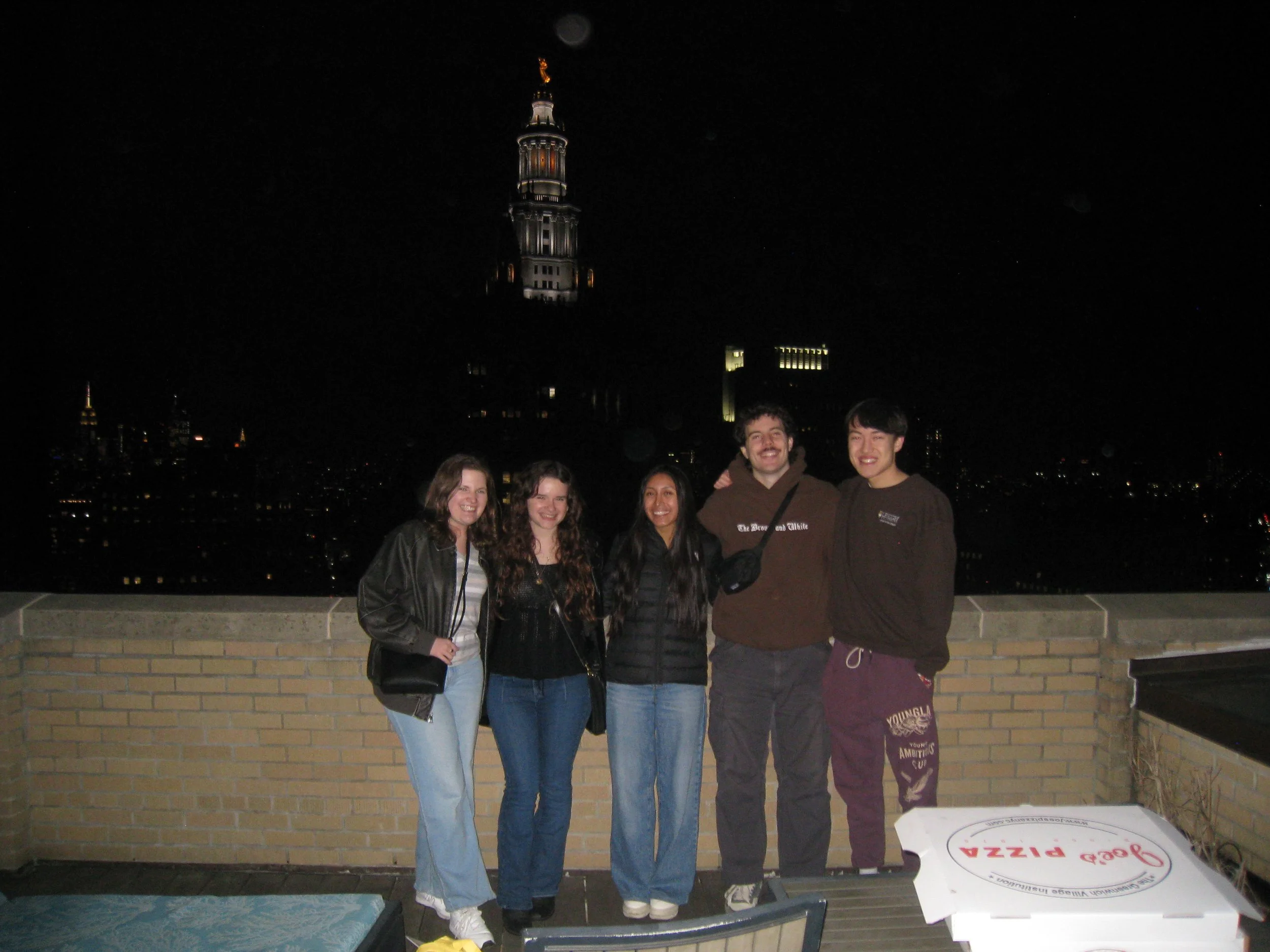Group of five young people standing on a rooftop at night with city skyline and the Empire State Building in the background.