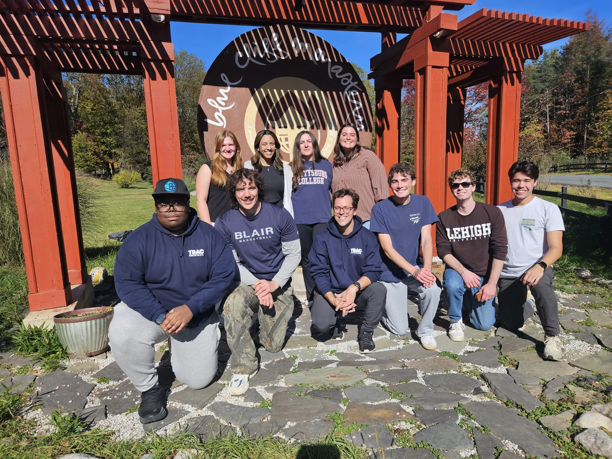 Group of ten young adults posing outdoors in front of a decorative wooden arch with a circular sign that reads "become a changemaker." They are standing on a stone pathway with trees and grass in the background on a sunny day.