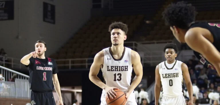 Lehigh University basketball players on the court during a game, with one player holding a basketball and preparing to shoot, and other players and spectators visible in the background.