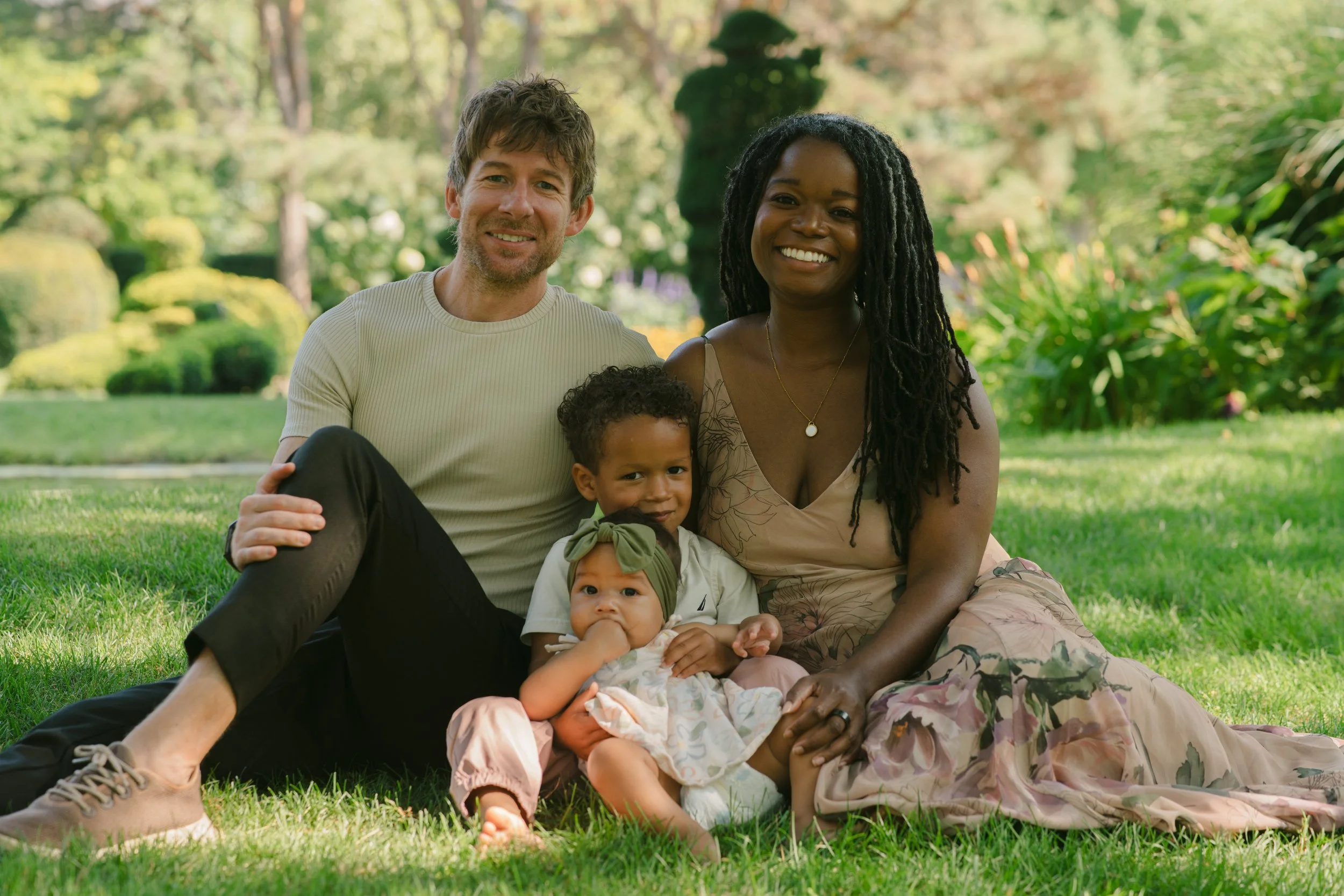 A happy multiracial family sitting on grass in a park, smiling at the camera. The group includes a man, woman, and two young children.