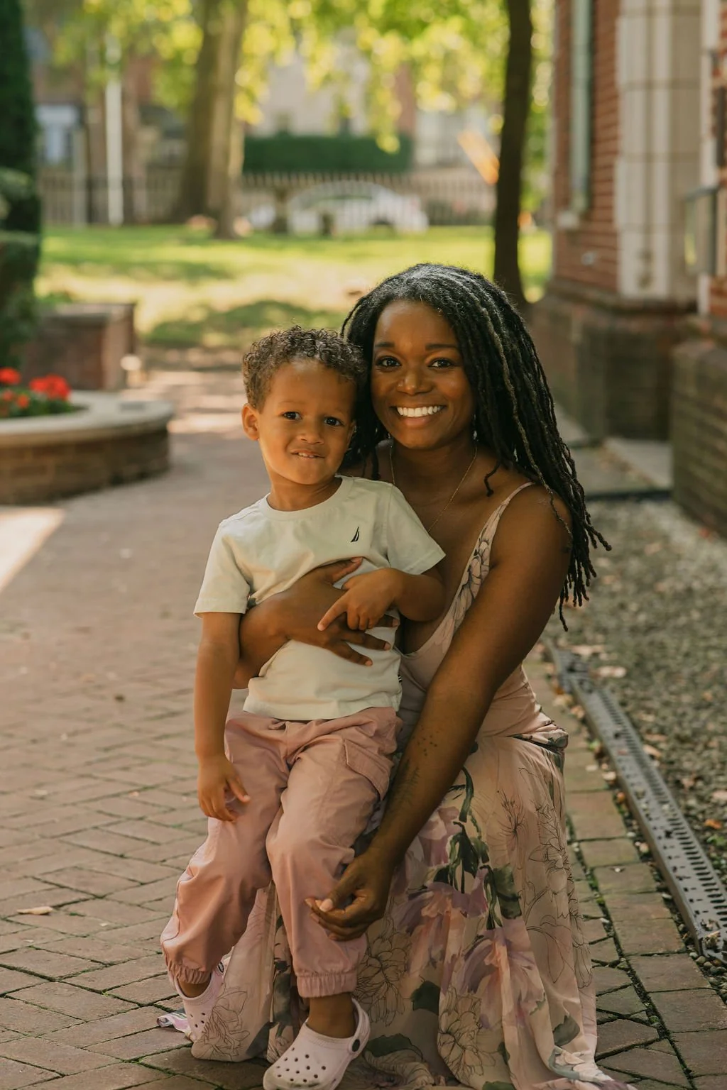 A woman with long, black, textured hair smiling while holding a young boy with curly hair outdoors on a brick sidewalk with trees and buildings in the background.