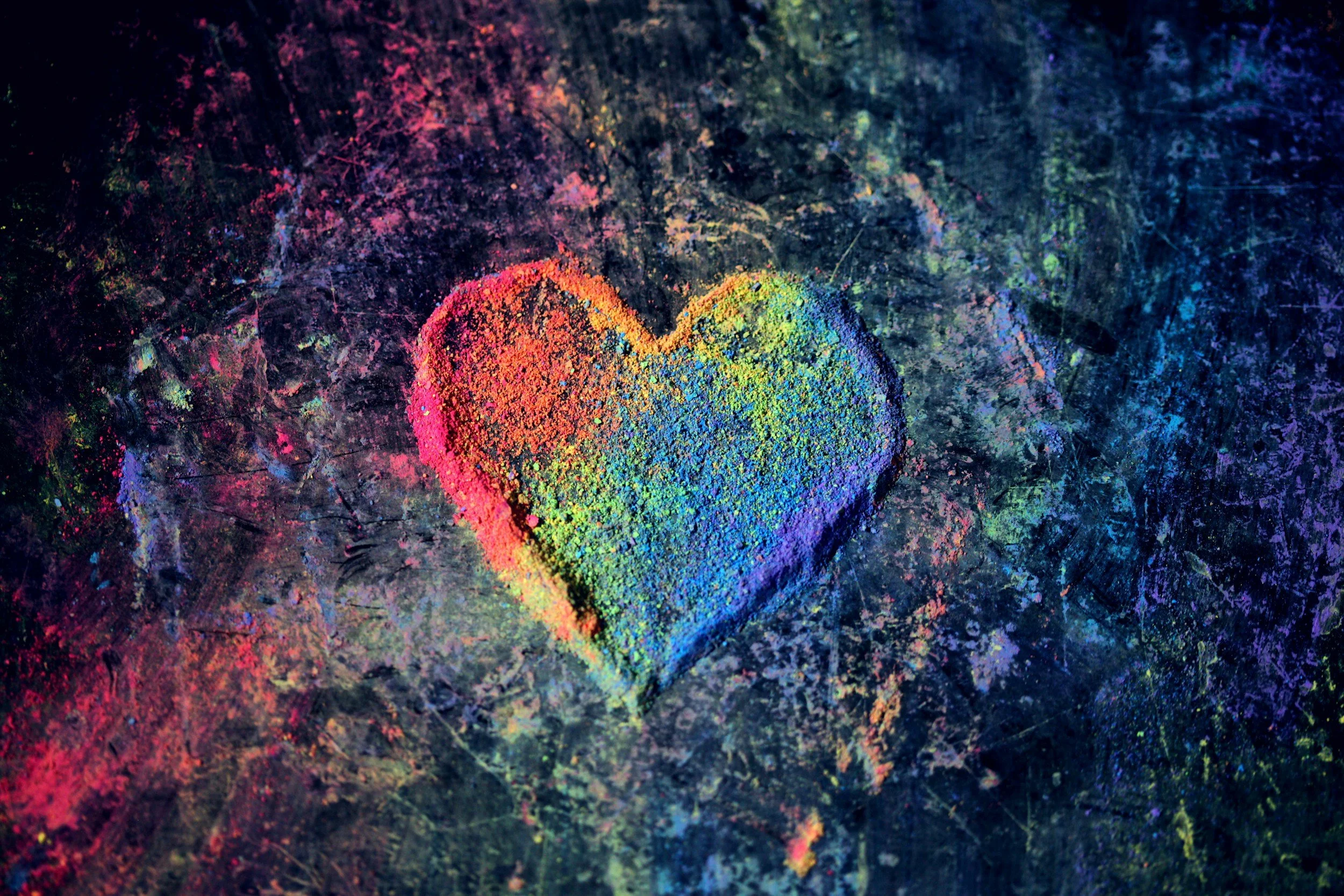 A heart shape made of rainbow-colored powder on a dark, textured surface.