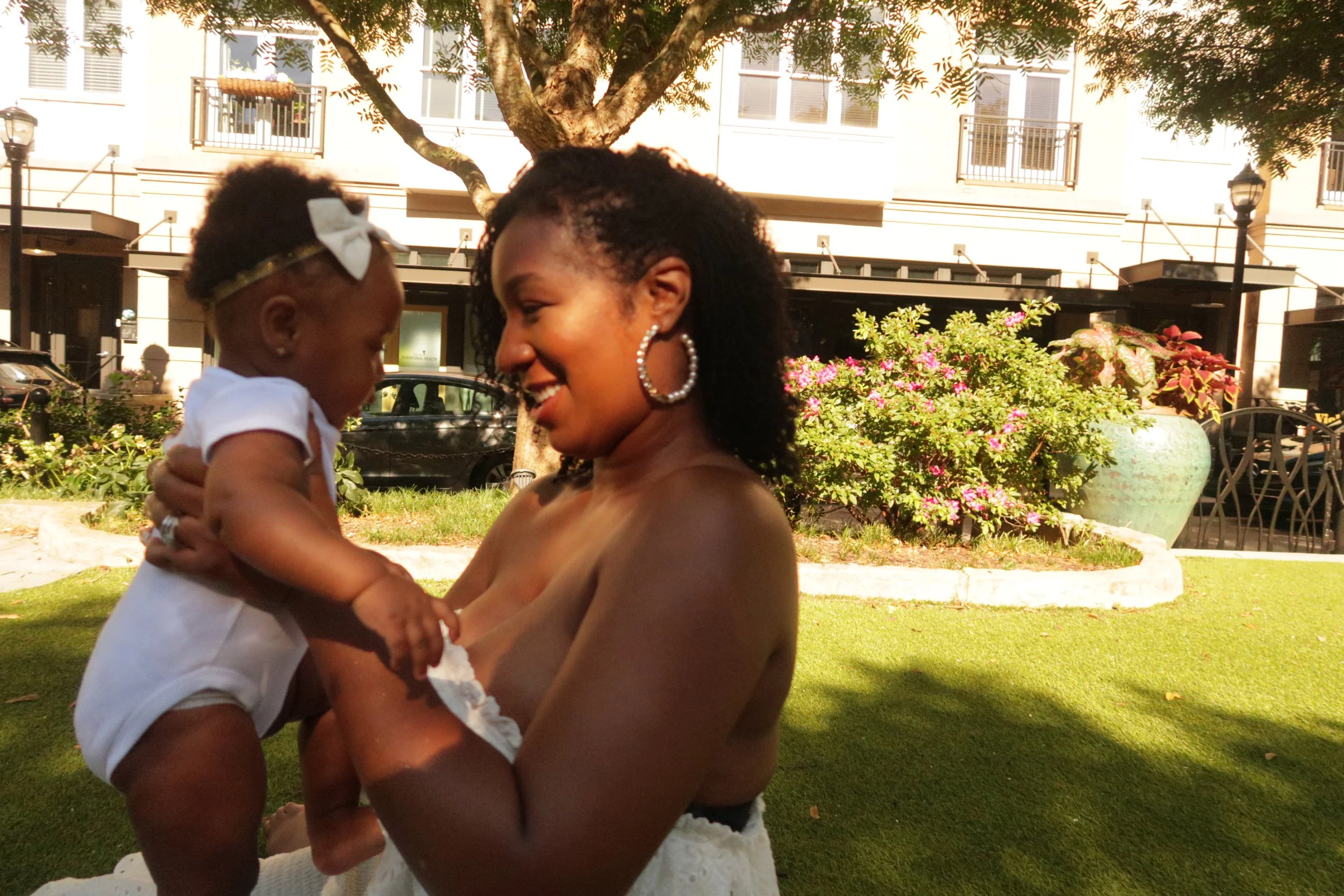 A woman with large hoop earrings holding a baby girl outside in a park area with green grass, trees, flowers, and buildings in the background.
