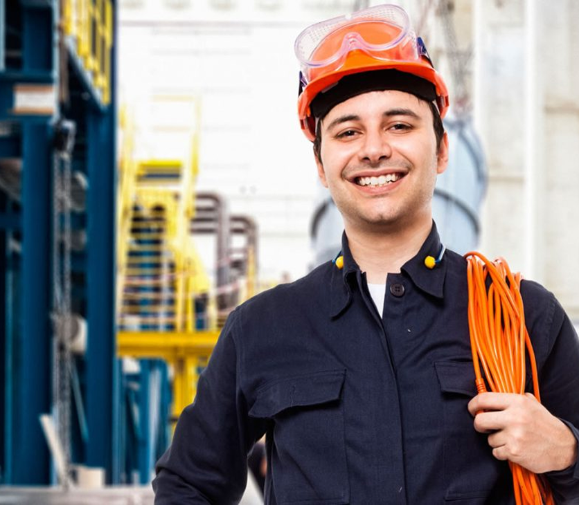 A young man smiling, wearing a safety helmet and goggles, holding an orange extension cord in a warehouse or industrial setting.