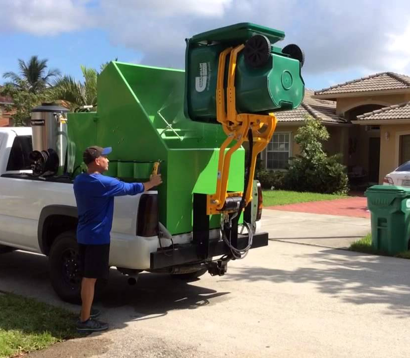 A man in a blue shirt and black shorts standing next to a pickup truck with a large green trash compactor mounted on the back.