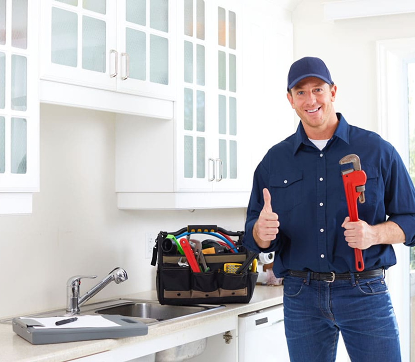 A man in a blue uniform and cap holding a red pipe wrench, smiling and giving a thumbs-up in a bright kitchen with white cabinets and a toolbox on the counter.