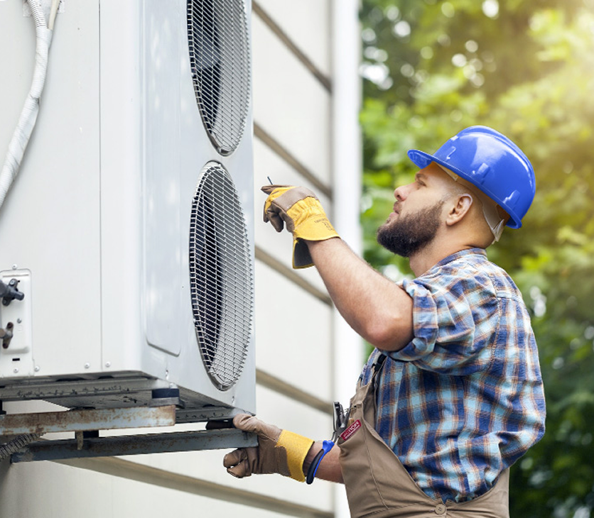 An HVAC technician wearing a blue helmet and yellow gloves installing or repairing an outdoor air conditioning unit on a house exterior.