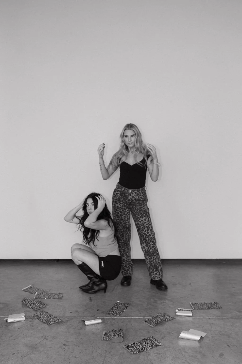 Two women posing in front of a plain wall with leopard print and plain clothing, with scattered leopard print papers on the floor.