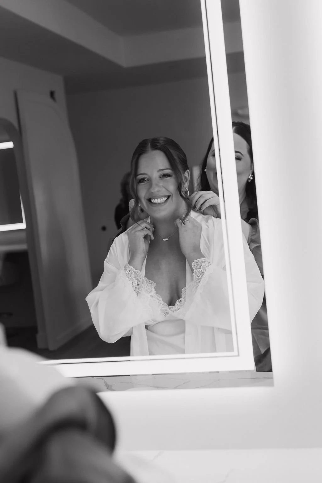 A woman in a satin robe smiling at her reflection in a mirror, assisted by another woman.