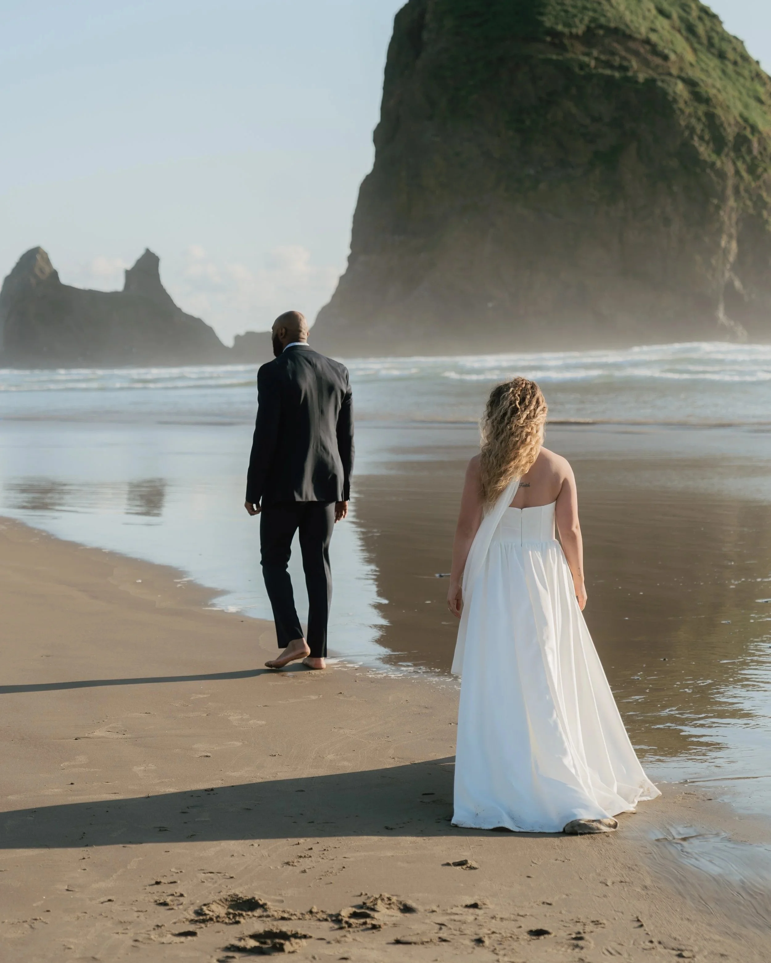 A man in a dark suit and a woman in a white dress walking along the beach near ocean waves with large cliffs in the background.