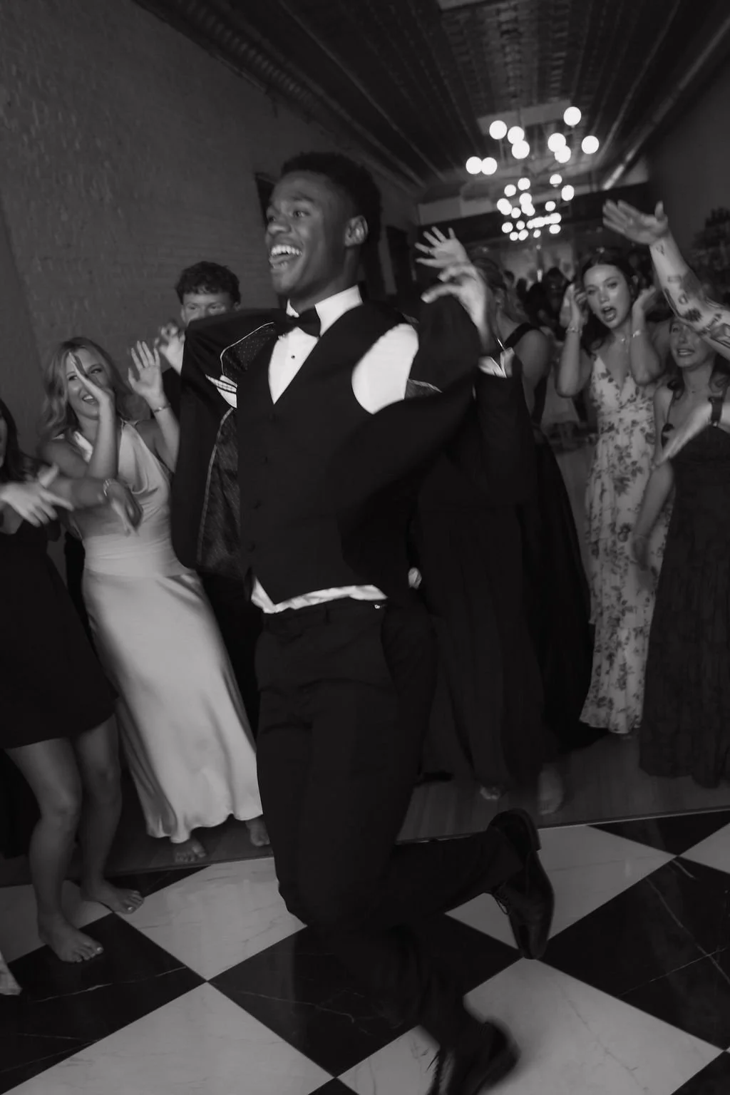 Black and white photo of a man in tuxedo dancing joyfully at a party with people in formal attire surrounding him, all appearing to be having fun.