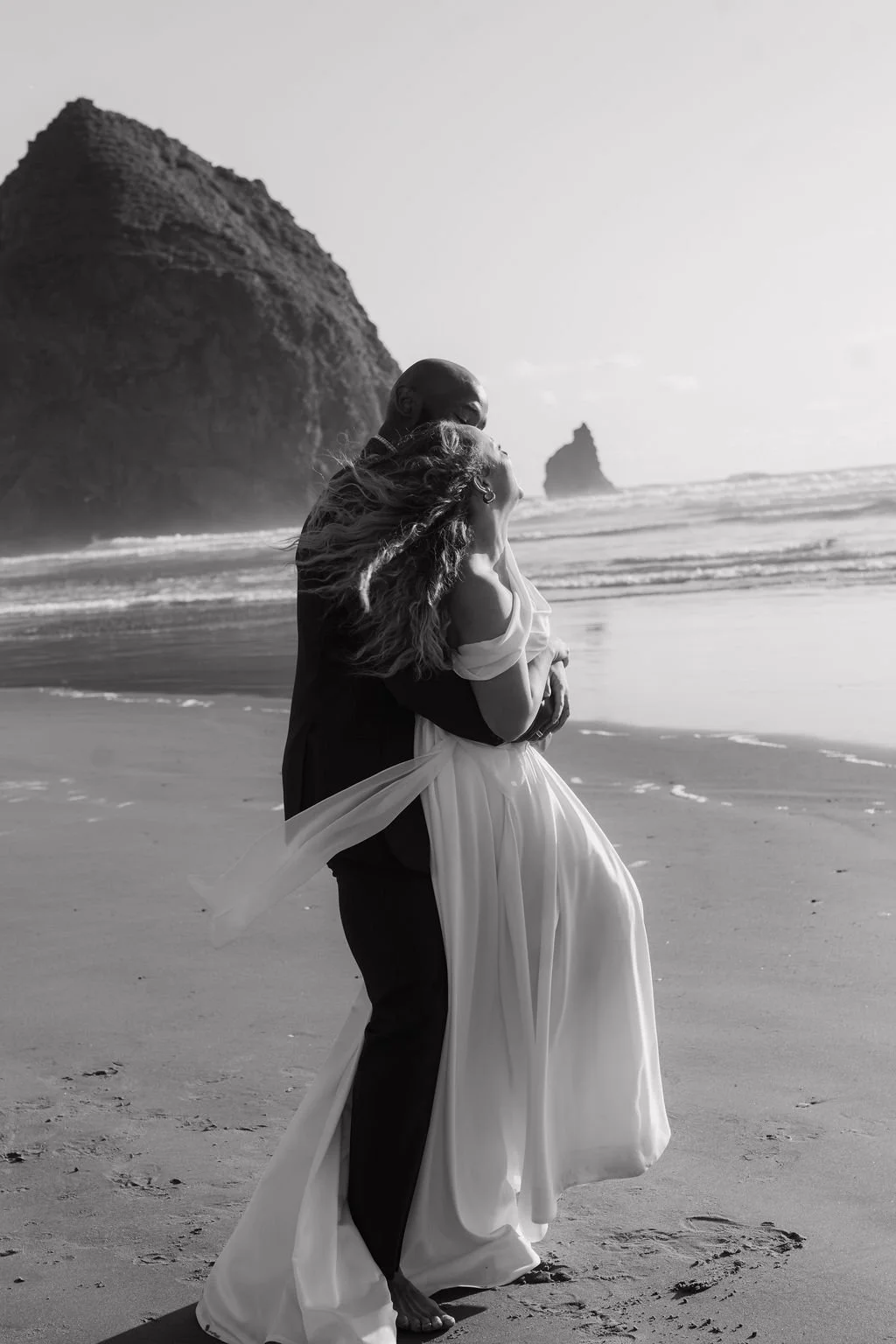 A black and white photo of a couple embracing on a beach, with large rock formations in the background and waves in the ocean behind them.