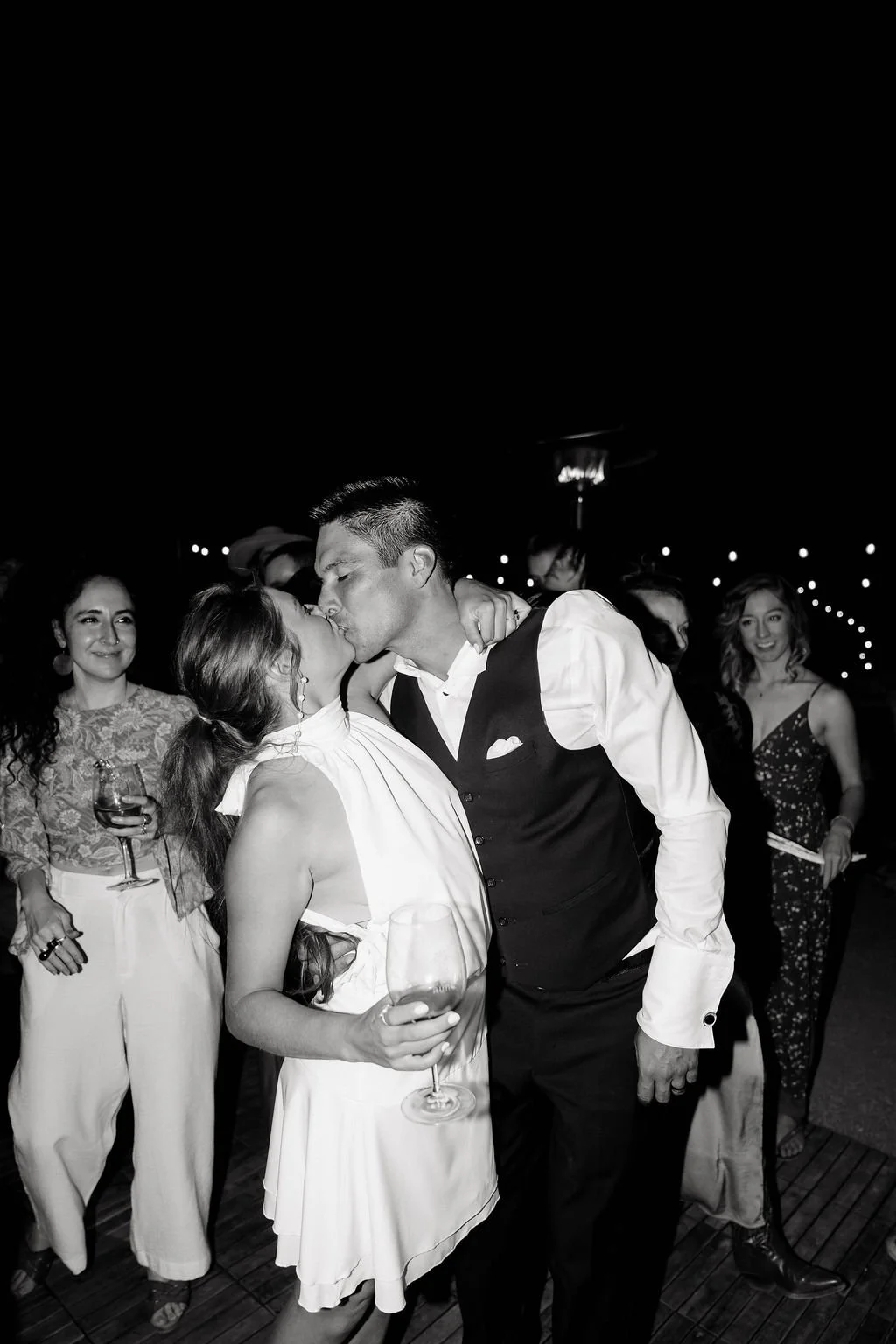 A couple sharing a kiss at a party, surrounded by friends holding drinks in a dimly lit venue.