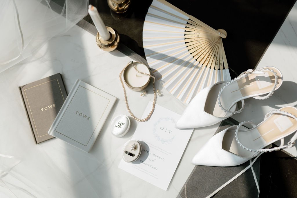 Flat lay of white high-heeled shoes, pearl jewelry, a decorative fan, a candle, two vow booklets, and an envelope on a white surface with sunlight and shadows.