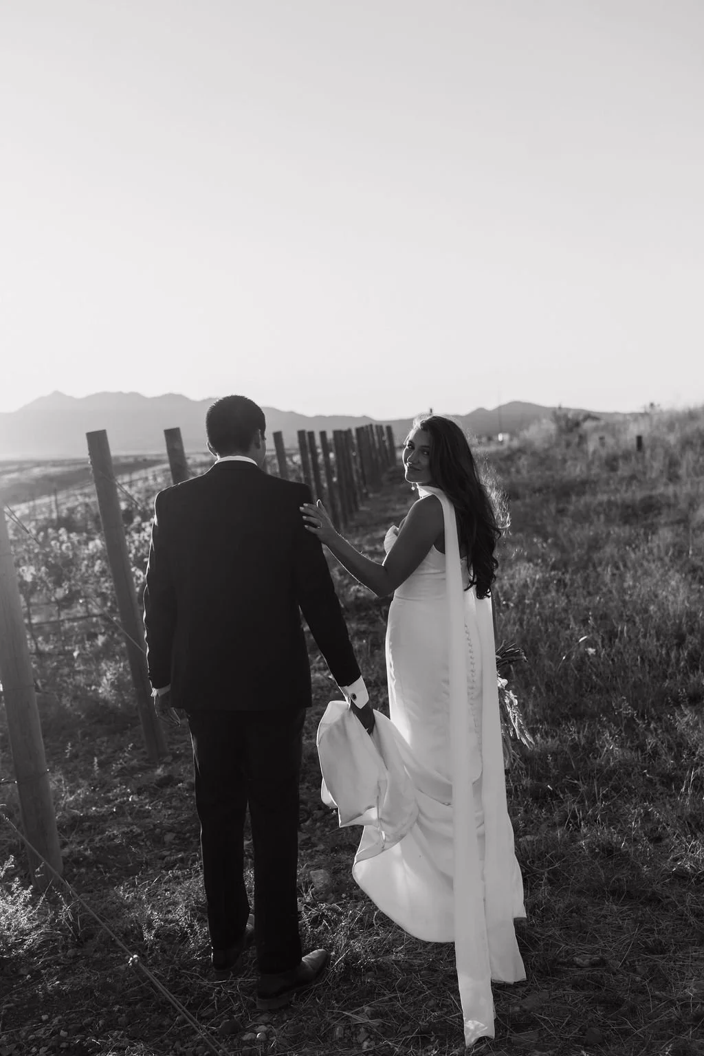A black and white photo of a couple in formal attire walking in a rural area with a fence and mountains in the background, the woman looking back and smiling.