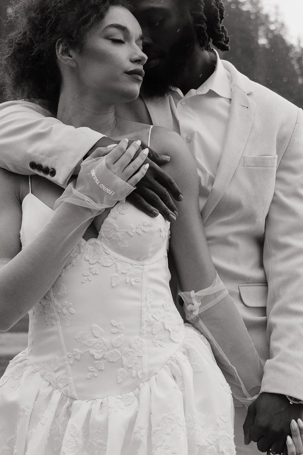 A black and white photo of a couple embracing, with the woman wearing a lace wedding dress and a glove with the text 'training trouble' on it, and the man in a light-colored suit, both with eyes closed.
