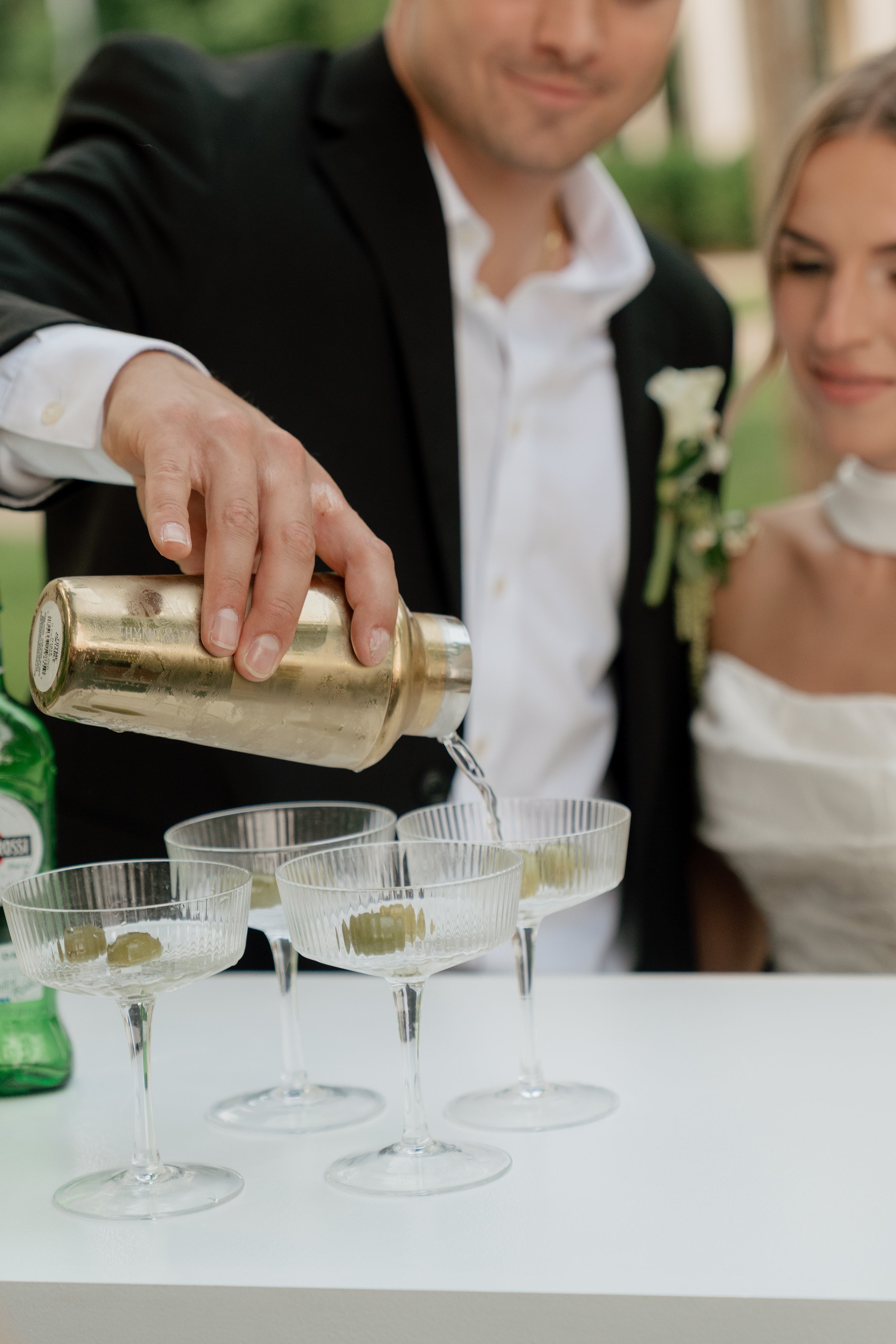 A man in a black tuxedo pours a clear liquid into glasses at a celebration, with a woman in a white dress standing nearby.