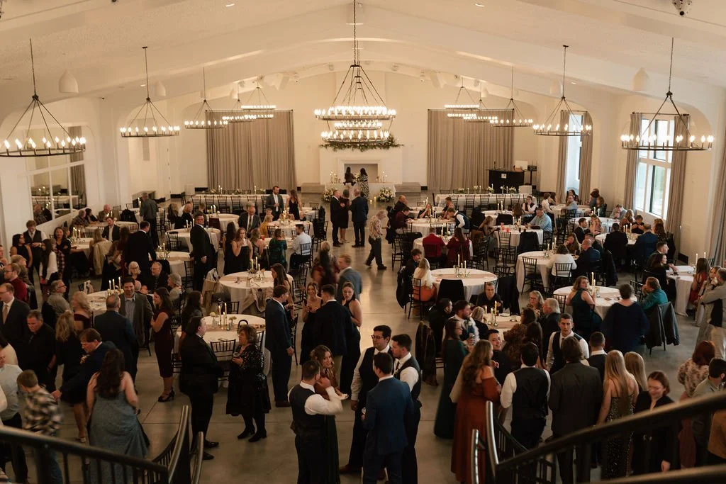 Wedding reception in a spacious, well-lit hall with guests mingling, sitting at round tables, and dancing.