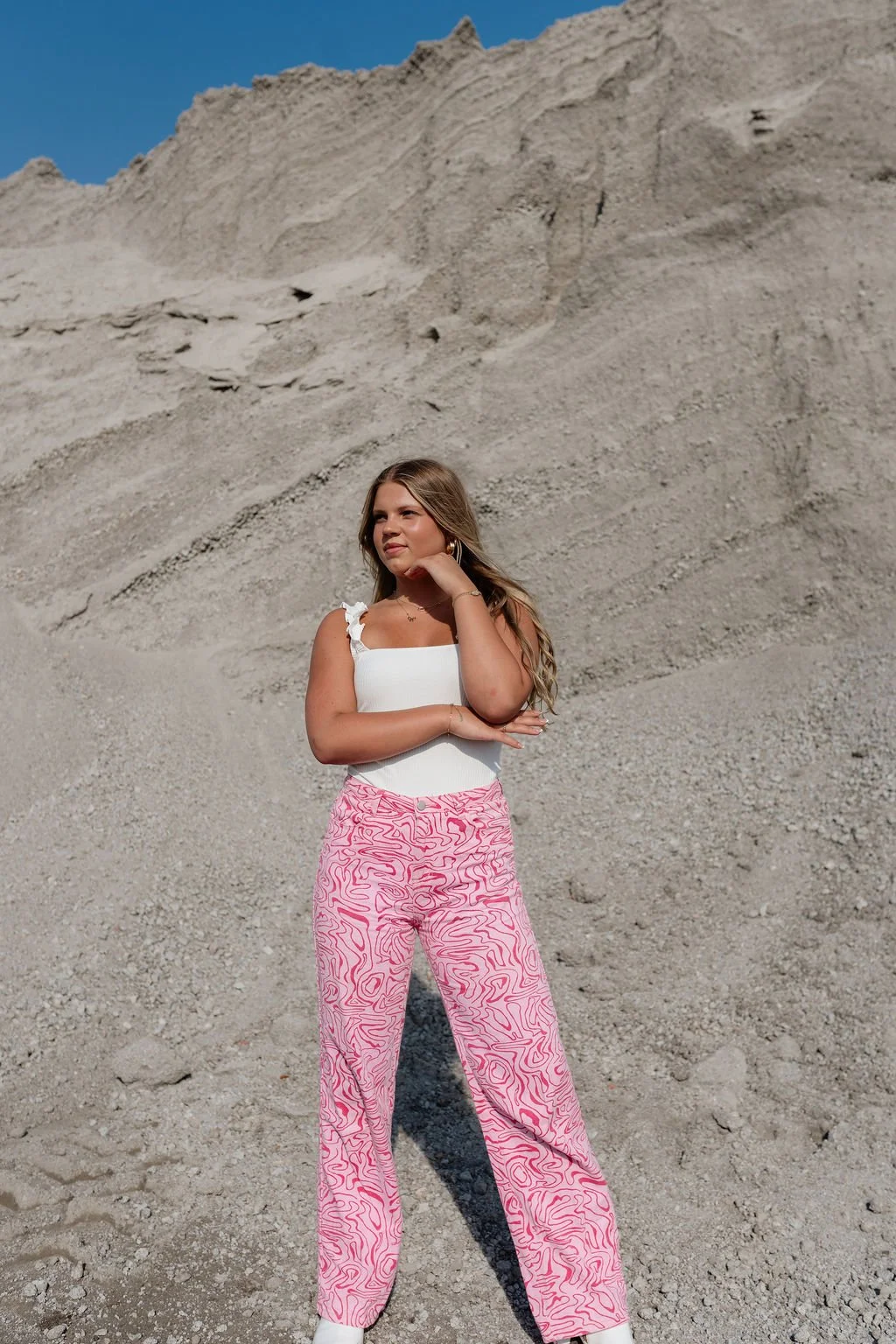A young woman with long, wavy hair stands in front of a rocky, sandy hill under a clear blue sky. She is wearing a white sleeveless top with ruffle straps and pink patterned wide-leg pants, with her right hand touching her neck and her left arm crossed over her stomach.