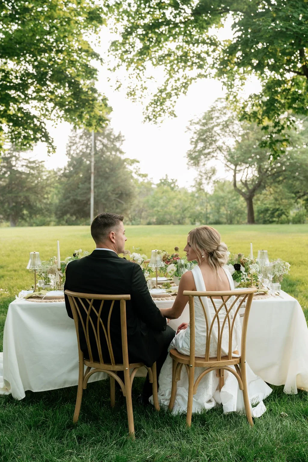 A bride and groom sit at a table outdoors in a lush green park, holding hands and facing each other during their wedding reception.