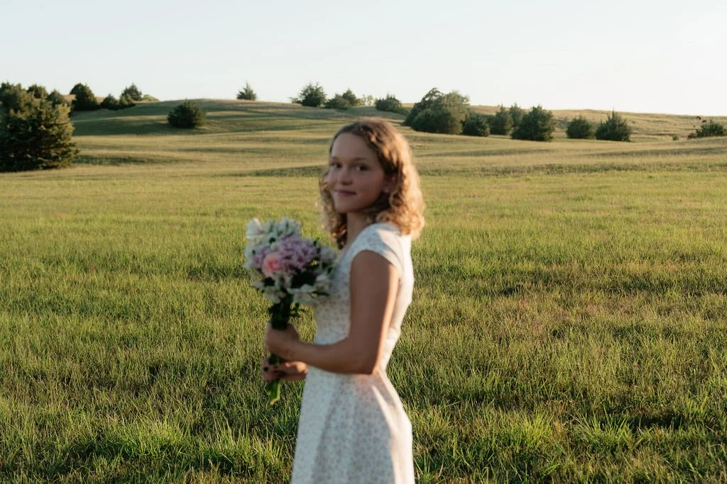 A young woman in a white dress holding a bouquet of flowers in a green field with trees in the background during sunset.
