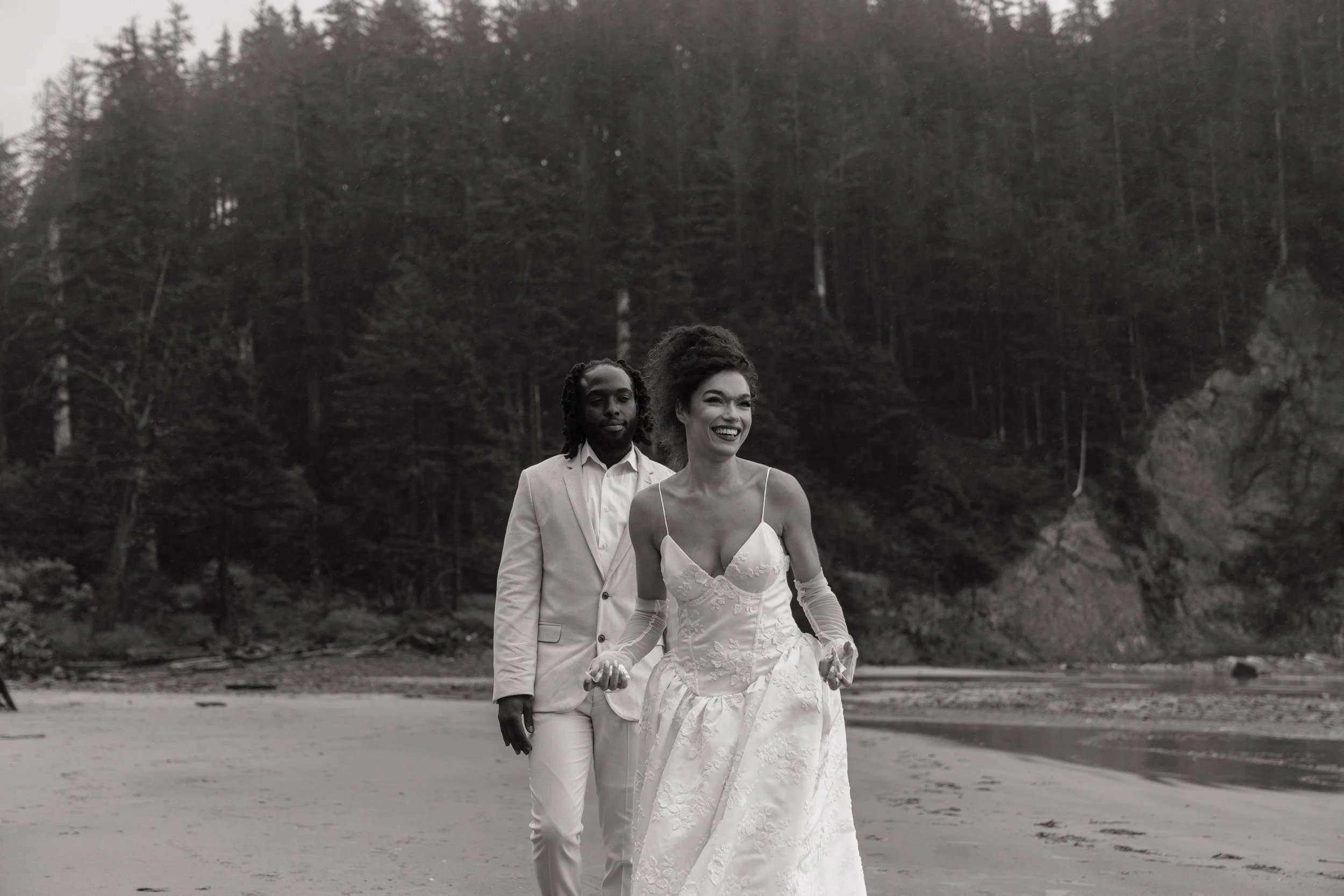 A woman in a wedding dress and a man in a suit walking on a beach with trees and cliffs in the background.