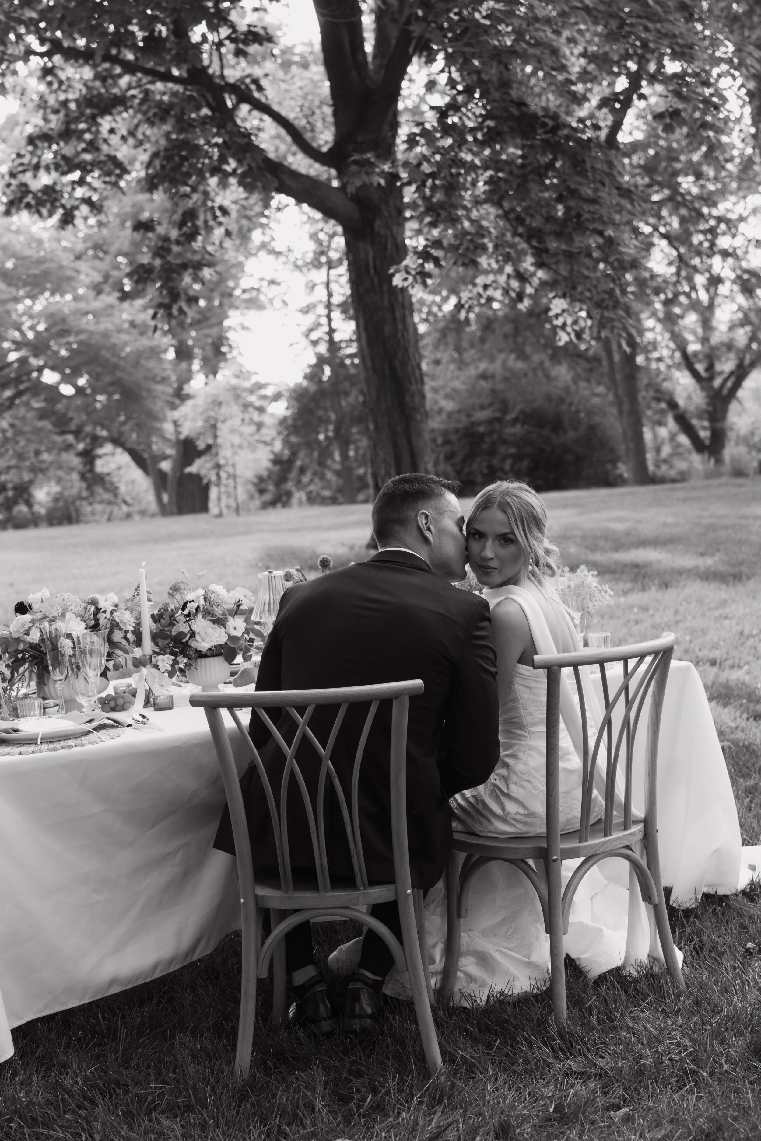 A black and white photograph of a bride and groom sitting at a decorated outdoor wedding table in a park or garden setting, with large trees and lush greenery in the background. The groom is kissing the bride on the cheek while she looks at the camera.