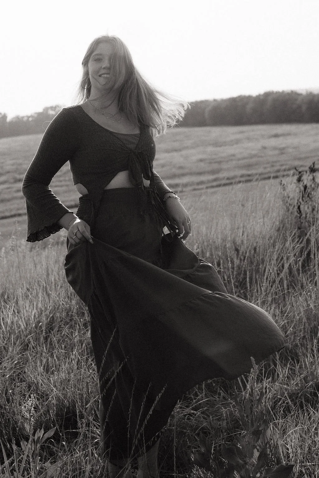 A woman standing in a grassy field, smiling and holding her dress with one hand, wearing a tied crop top, jewelry, and a long skirt, with trees and open land in the background, in black and white.