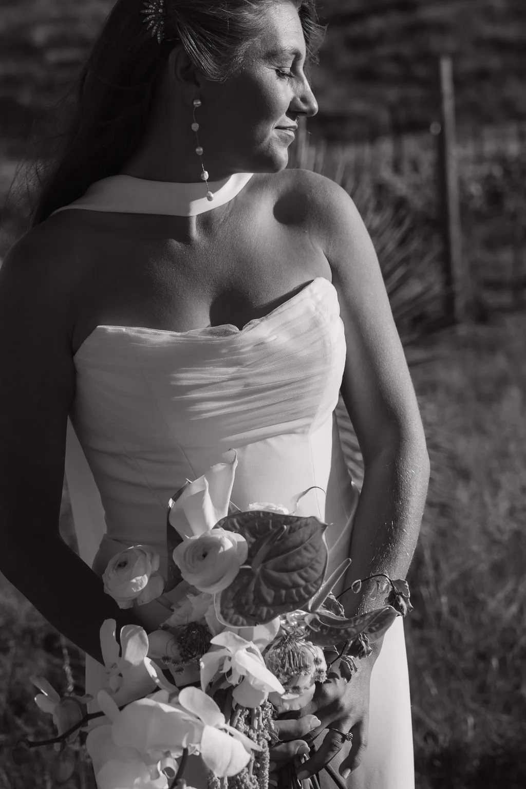 A woman in a strapless white dress holding a bouquet of flowers, standing outdoors with her eyes closed.