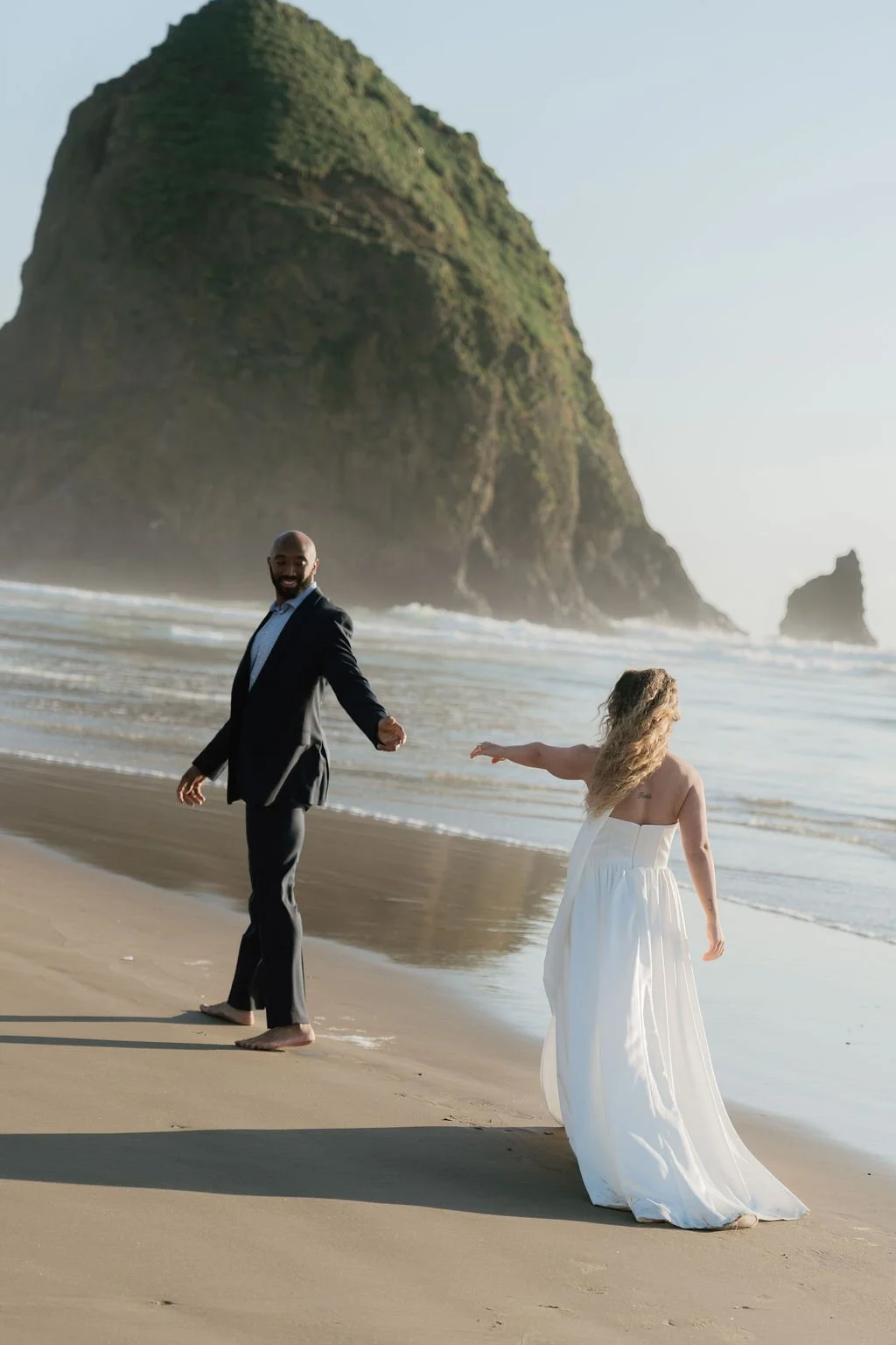 A man in a black suit and barefoot, holding hands with a woman in a white dress, walking on a sandy beach with a large rocky island in the background.