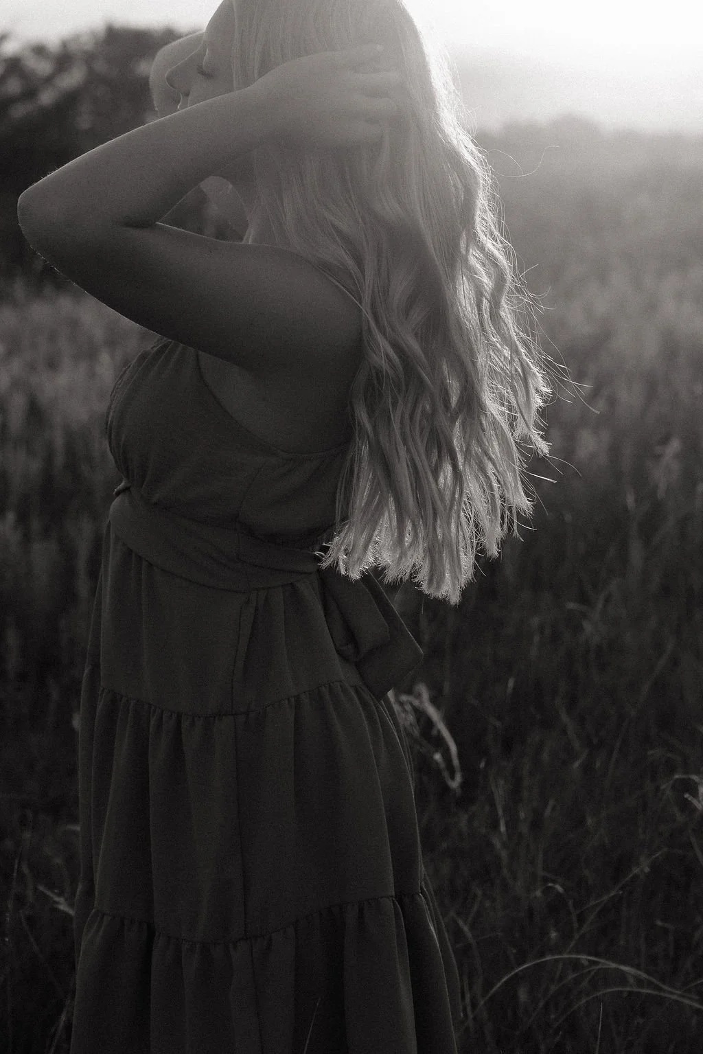 A woman with long, wavy hair standing outdoors in a field at sunset, with her arms raised, touching her hair.