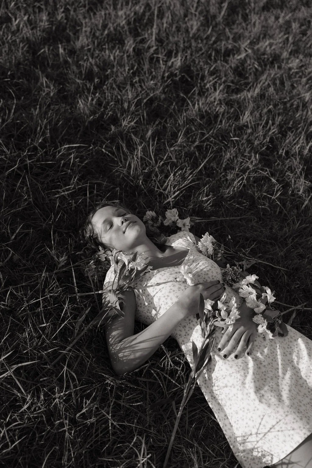 A young woman lying on her back in grass with closed eyes, holding flowers on her chest and around her, wearing a light-colored dress with a floral pattern.