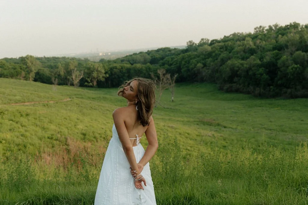 A woman in a white strapless dress with her eyes closed standing in a green field with hills and trees.