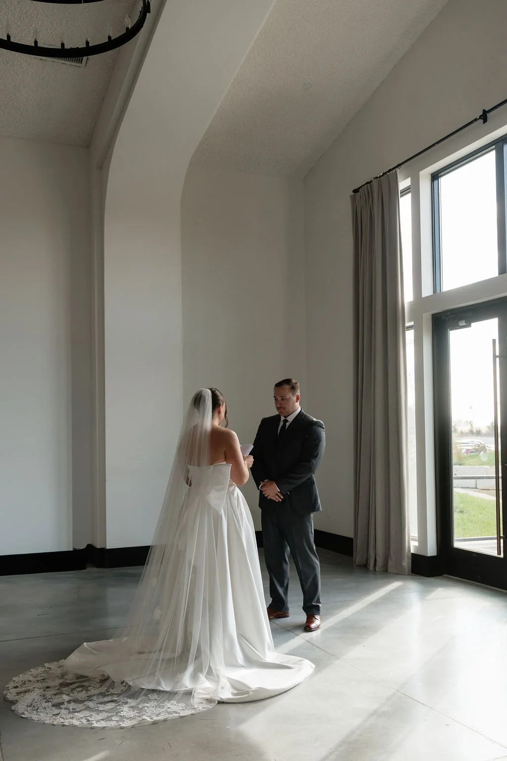 A bride and groom standing face to face in a bright room with large windows and white walls, during their wedding ceremony.