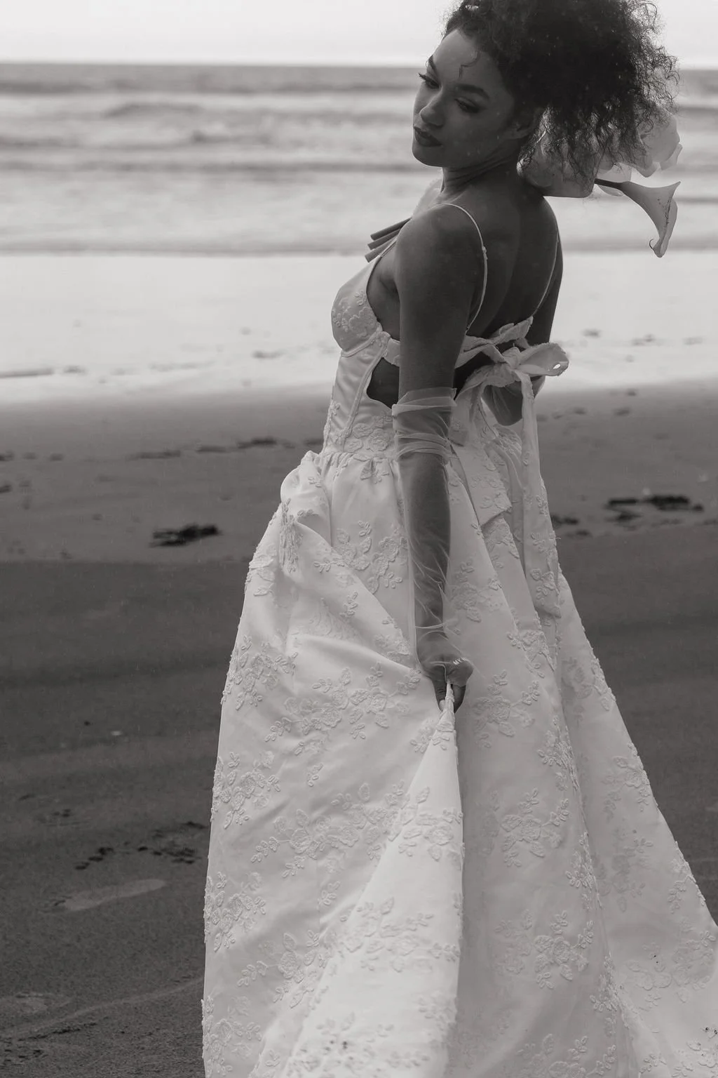 A woman in a wedding dress holding a flower walks on the beach.