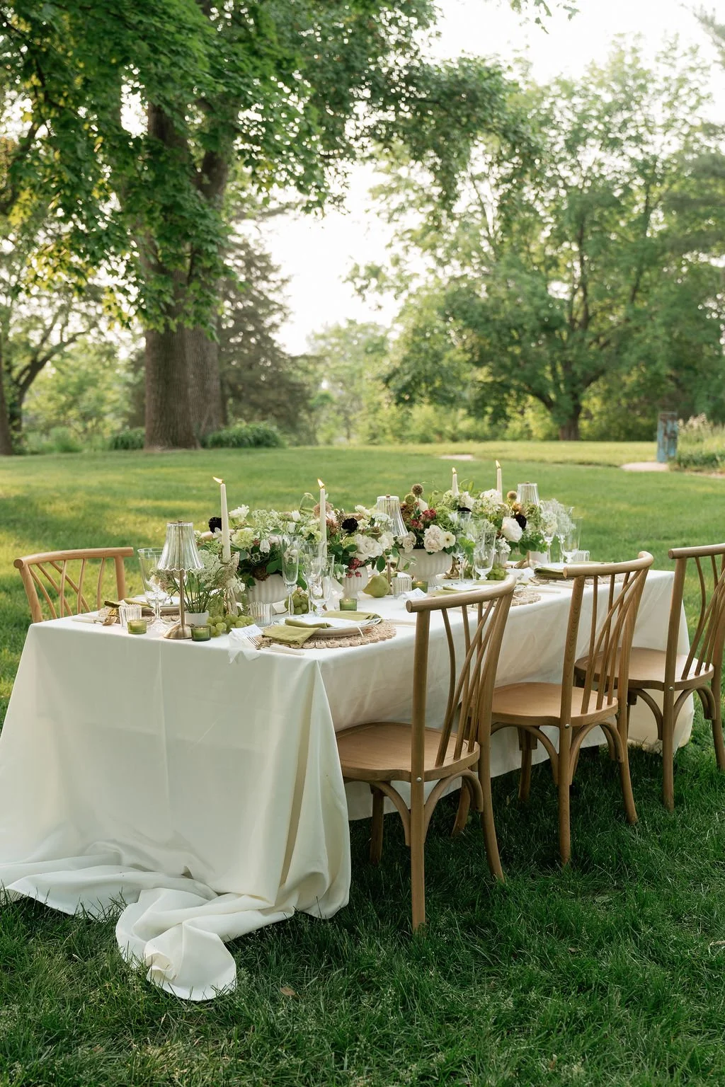 An outdoor dining table decorated with white flowers, candles, and glassware, set in a lush green park with large trees in the background.