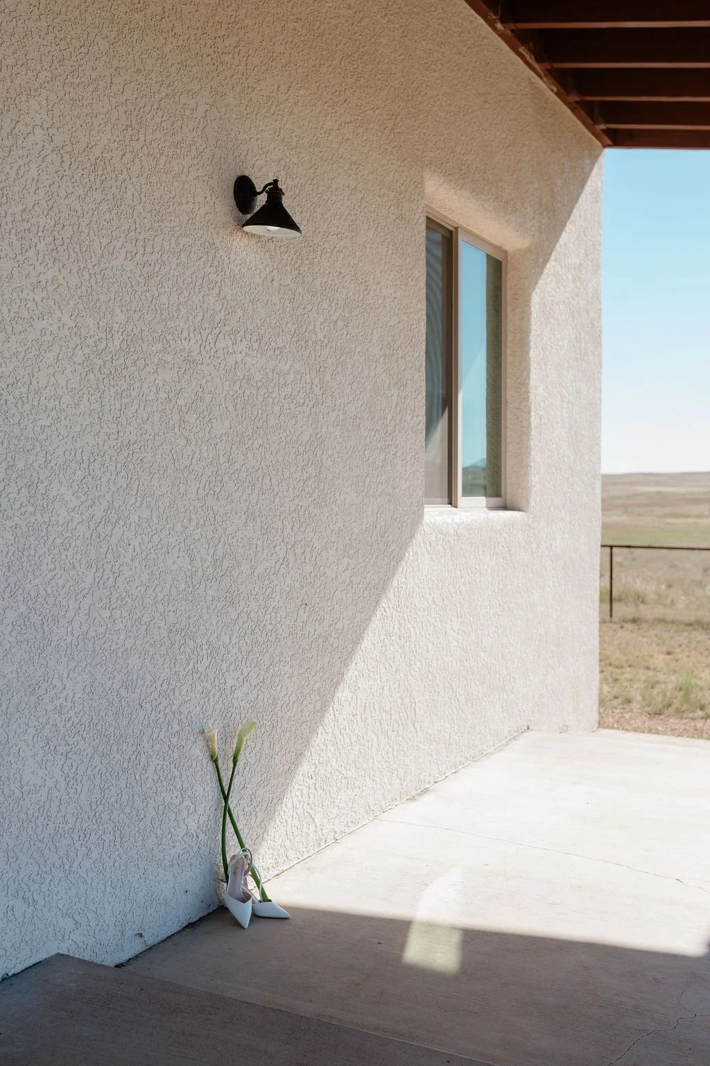White higher heels with lilies leaned against a textured cream-colored wall on a porch. A black outdoor wall light is mounted above, and there is a window with a view of the outdoor landscape.