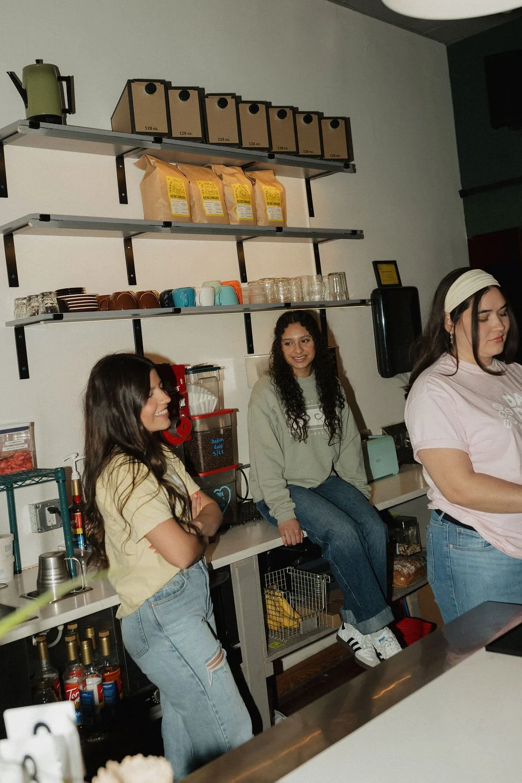Three women in a coffee shop, engaging in conversation, with shelves of coffee bags, cups, and kitchen supplies behind them.
