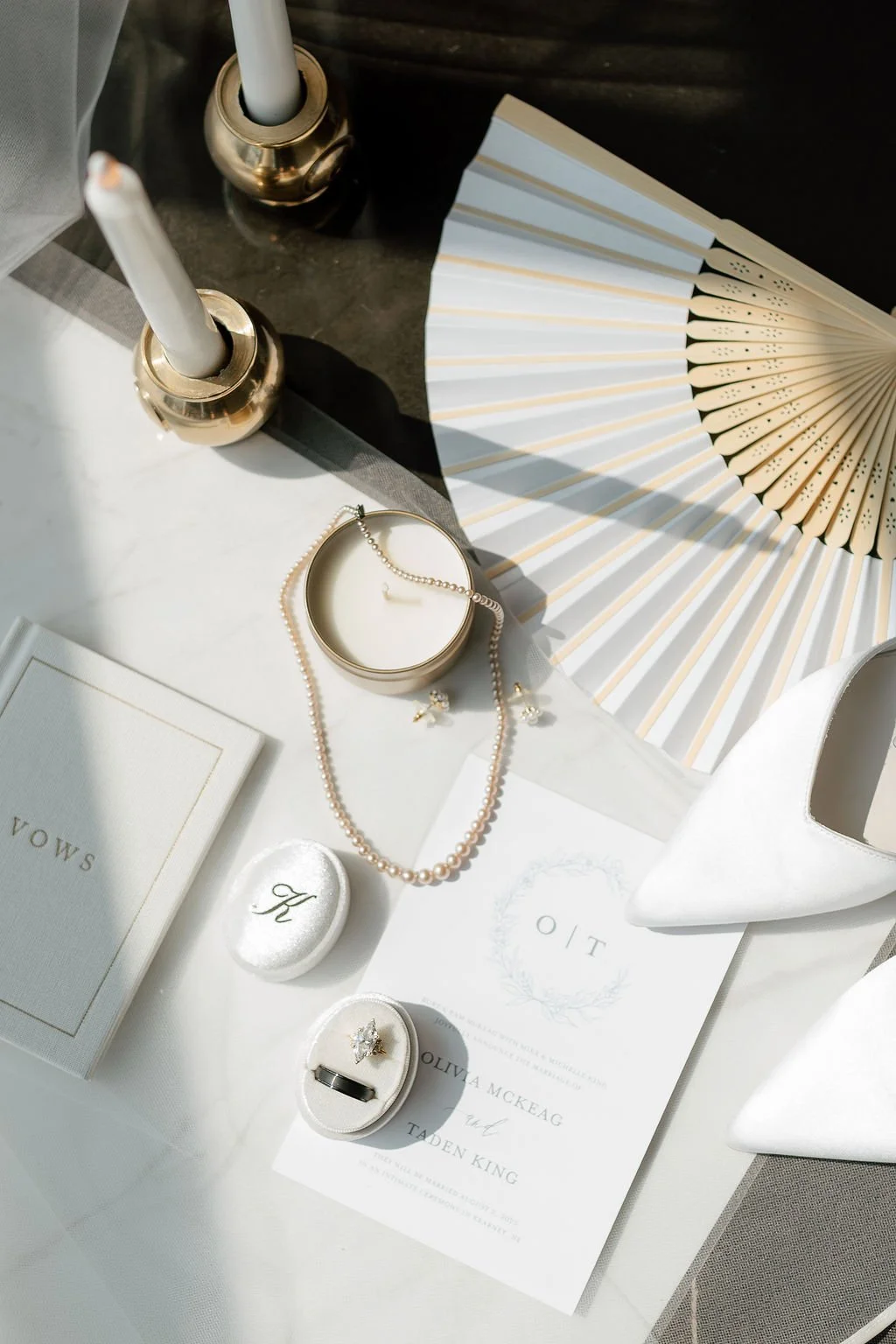 Wedding invitation, pearl necklace, pearl earrings, rings, candle, fan, white shoes, candles, books, and decorative items on a table.