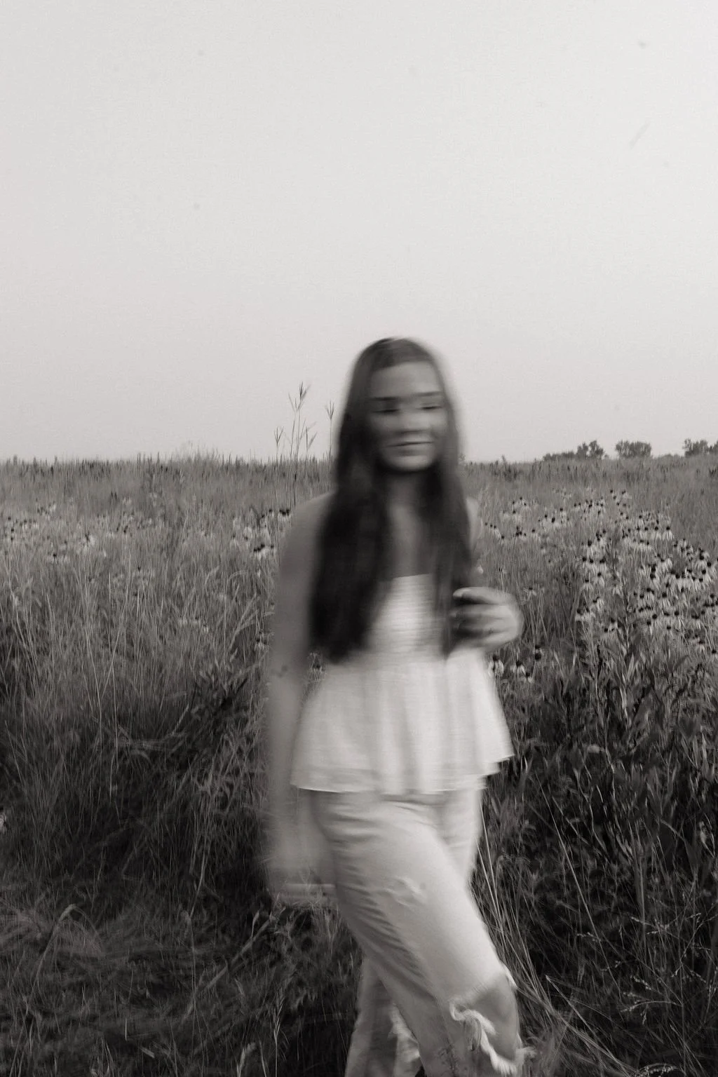 A woman with long hair standing in a field of tall grass and wildflowers, smiling and looking at the camera, blurred in black and white.