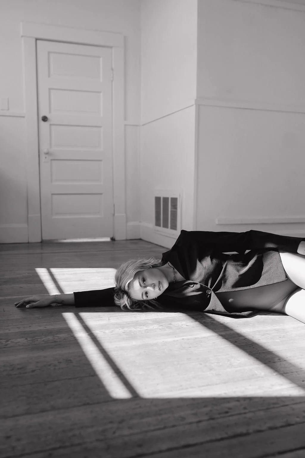 A woman lying on the wooden floor of a room, illuminated by sunlight through a window, with a closed door and a vent on the wall behind her.