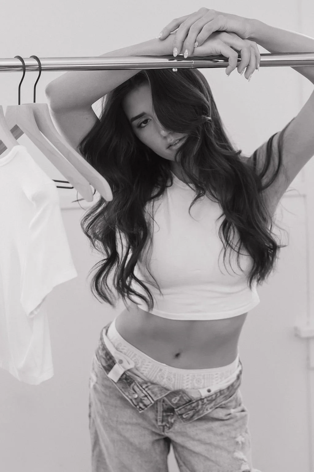 Black and white photo of a young woman with long, wavy hair, posing with her arms resting on a clothing rack with hangers, wearing a cropped top and jeans.