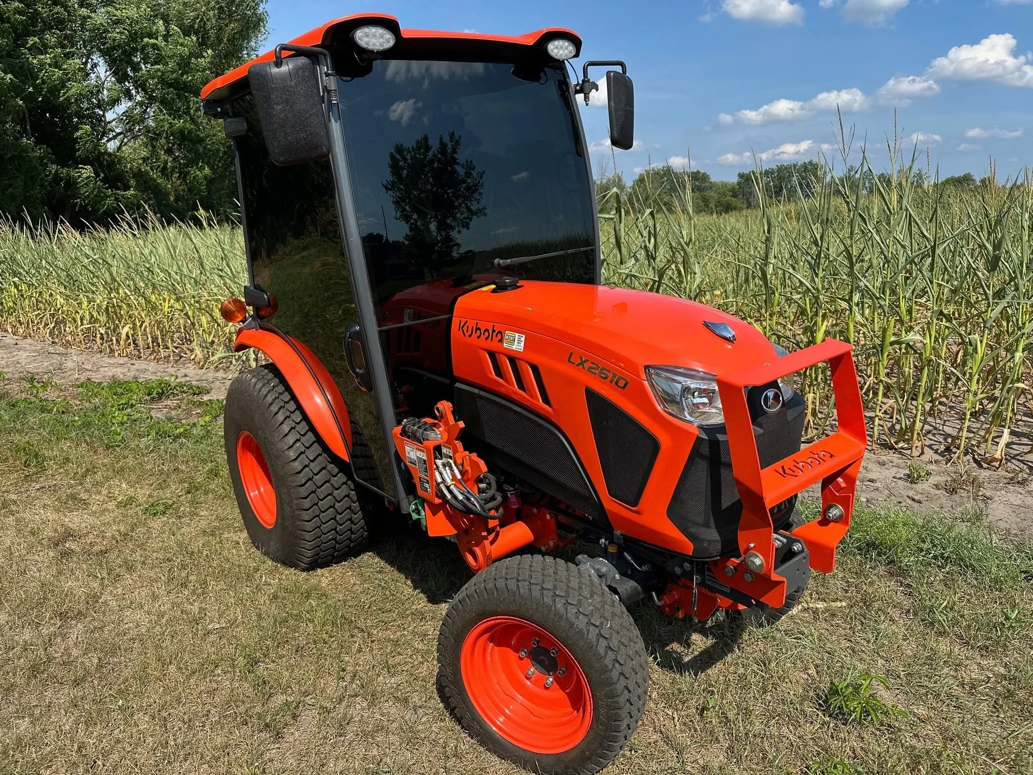 Orange Kubota tractor on a grassy field with tall green plants in the background and a blue sky with scattered clouds.