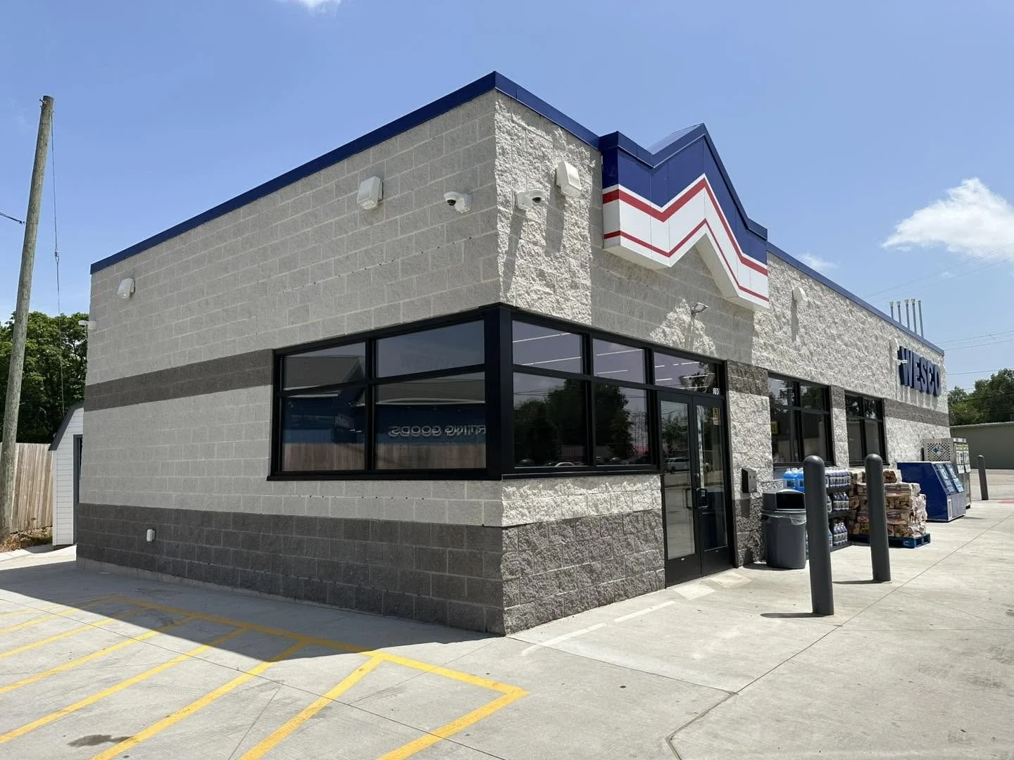 Exterior of an ESCO convenience store with large glass windows, a small parking lot with yellow lines, and a blue sky.