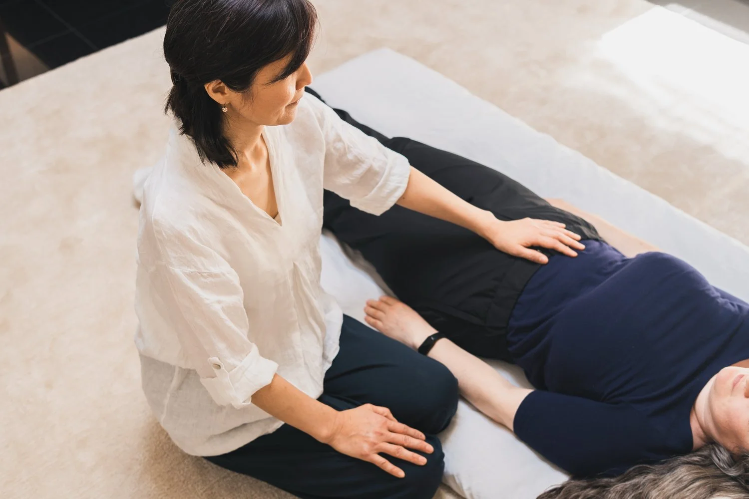 A woman practicing Reiki on a person lying face down on a massage table. She is kneeling on the floor with her hand gently placed on the person's back.